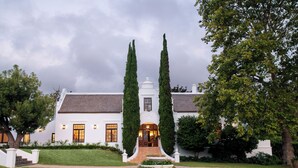 A stately white building with a symmetrical facade, a central entrance with steps, and tall, slender trees flanking the entrance. - Avontuur Manor House (Stellenbosch)