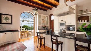 A kitchen with wooden beams, white cabinets, a large island, bar stools, and a view through a glass door. - Avontuur Manor House (Stellenbosch)