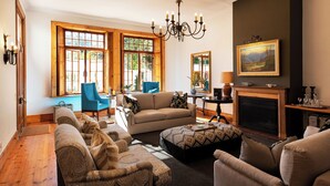 A living room with a fireplace, a chandelier, and a large window with a view of trees. - Avontuur Manor House (Stellenbosch)