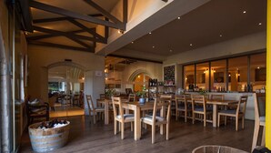 A spacious dining area with wooden tables and chairs, a bar area with a chalkboard menu, and a view of the outdoor garden through large windows. - Avontuur Manor House (Stellenbosch)