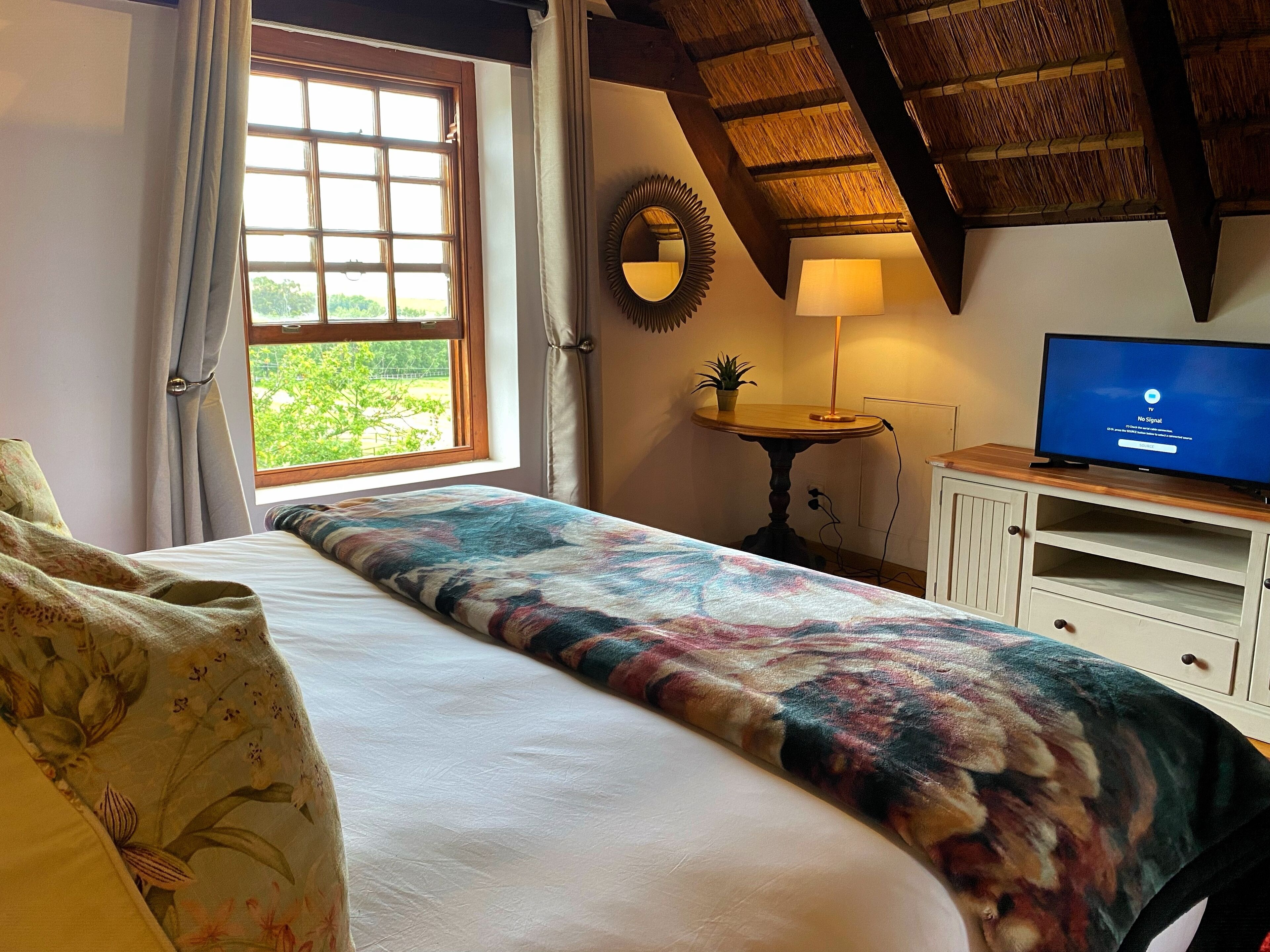A bedroom with a bed, a window with curtains, a bedside table with a lamp, a TV on a wooden cabinet, and a ceiling with exposed beams.
