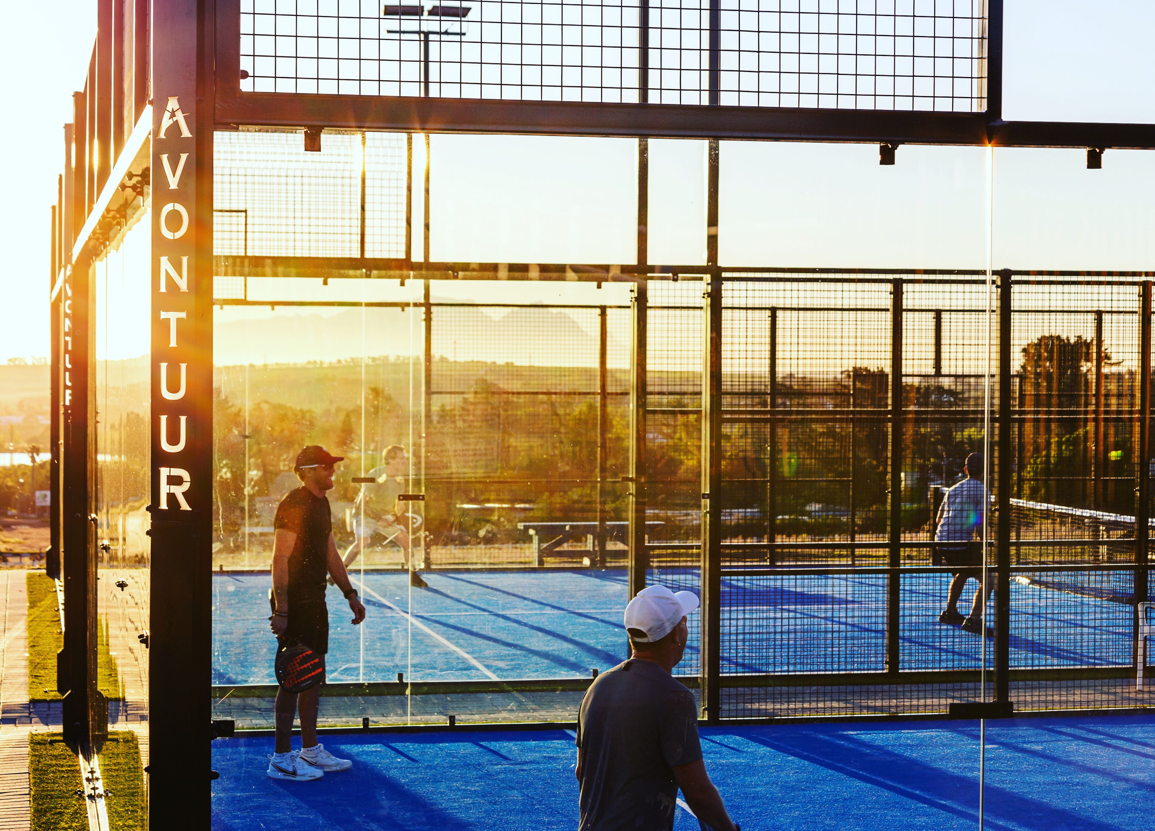 A tennis court with a net, surrounded by a fence, and a sign reading "AVONTUR".