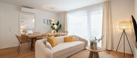 Un salon moderne comprenant un canapé, une table à manger et des chaises, une petite table d’appoint avec une plante, et une grande fenêtre avec des rideaux.