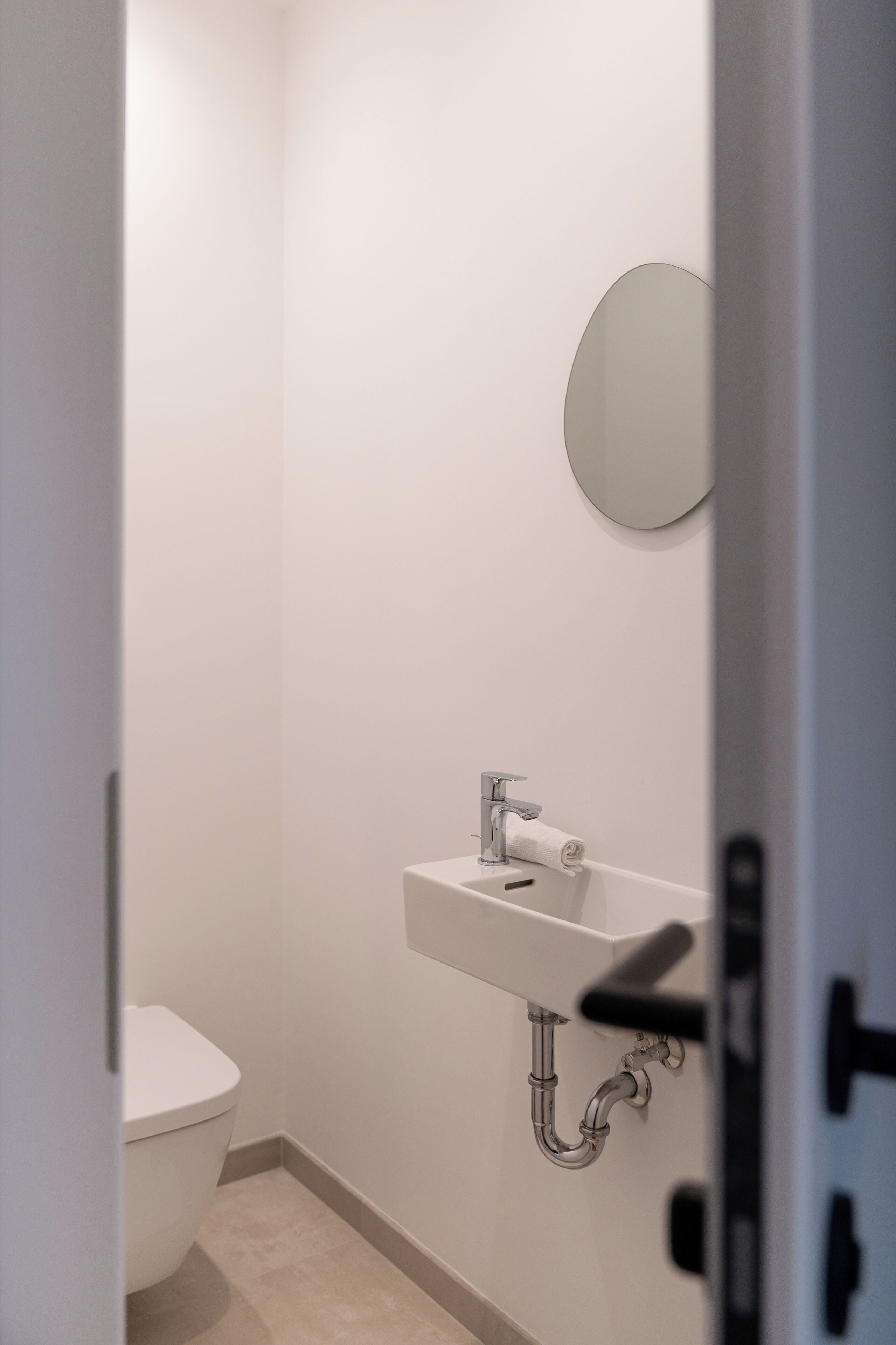 Et lille, moderne badeværelse med et hvidt toilet, en vask med spejl og et håndklæde hængende på væggen.