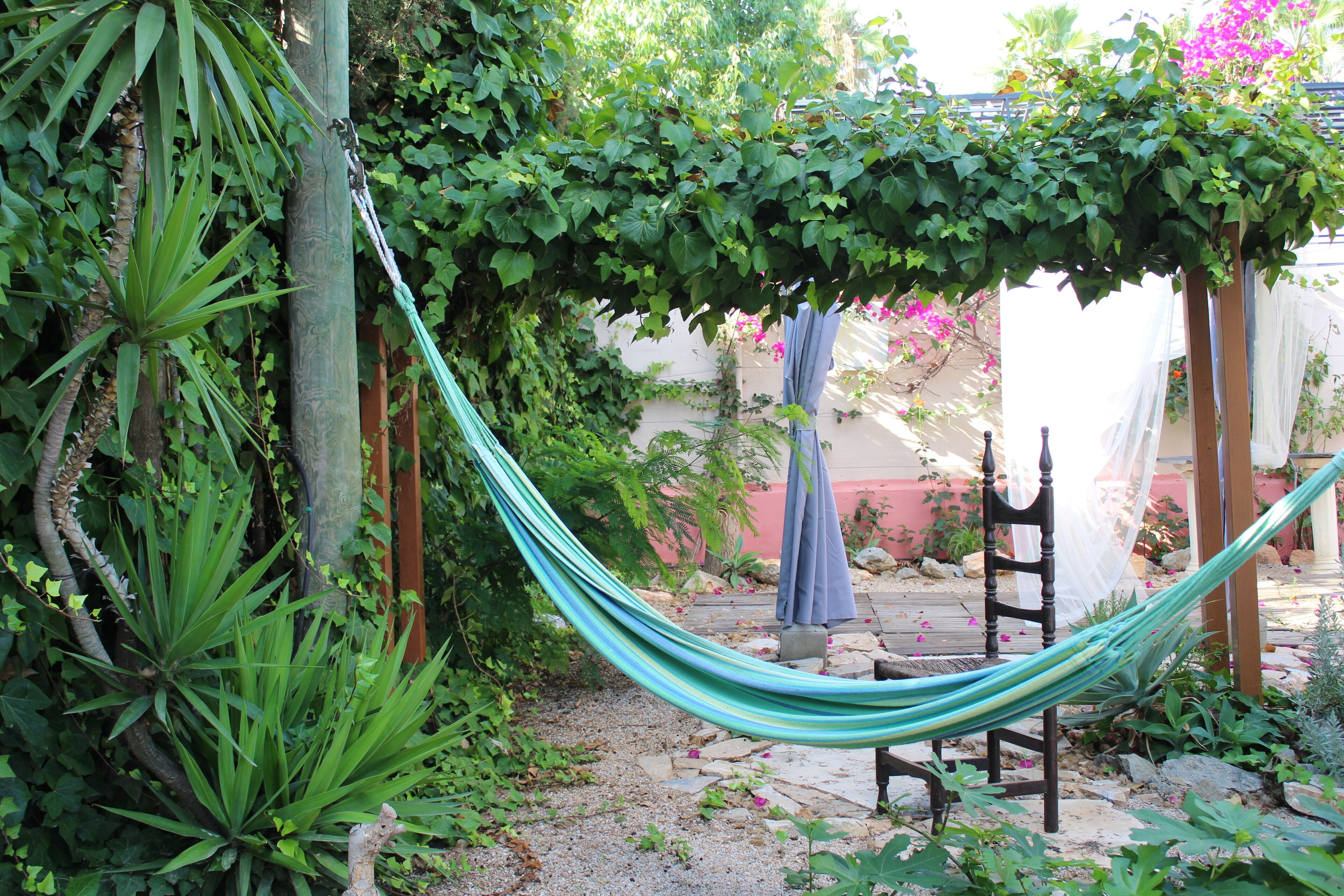 Un hamac est suspendu entre deux poteaux en bois dans un jardin où la végétation luxuriante côtoie un sentier pavé de pierres.