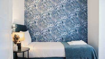 Un dormitorio con papel tapiz estampado, una cama con ropa de cama blanca y azul, una mesita de noche con una lámpara y un jarrón con flores.