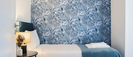 Un dormitorio con papel tapiz estampado, una cama con ropa de cama blanca y azul, una mesita de noche con una lámpara y un jarrón con flores.