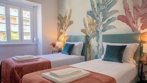 A hotel room with two beds, each with a white comforter and a folded white towel. There is a window with blinds, a nightstand with a lamp, and a wall with a large plant mural. - Succeed Campo Pequeno Suites (Lisbon)