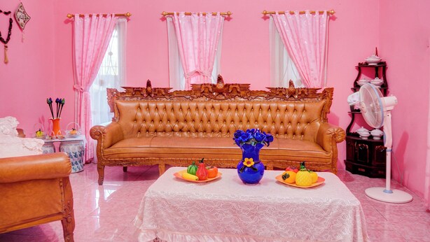 Una habitación con pared rosa, sofá marrón, mesa blanca con frutas y un jarrón azul, y un ventilador.