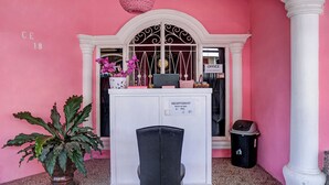 Un mostrador de recepción con un letrero que indica la oficina del recepcionista, una planta en maceta, un basurero y una silla frente al mostrador.