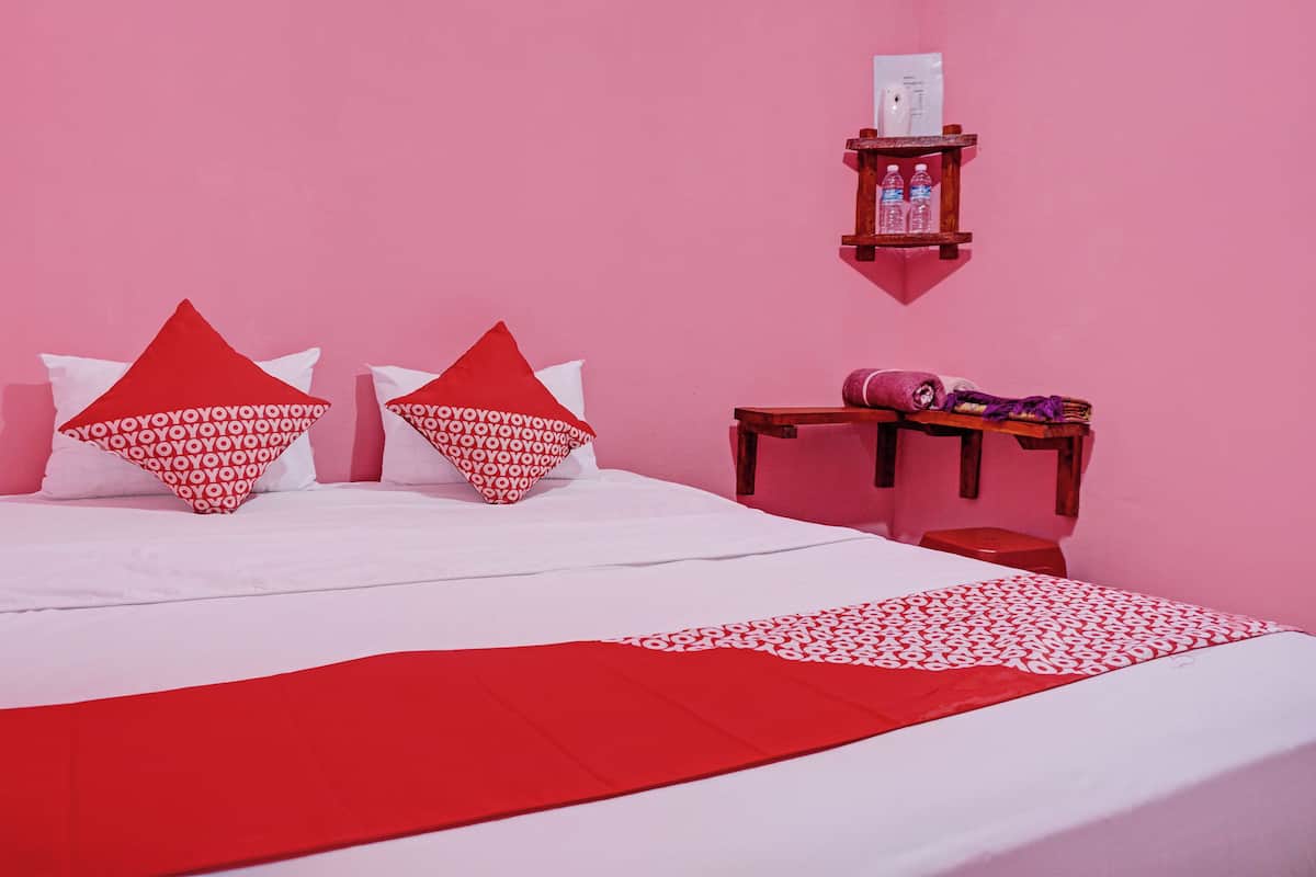 A bed with red and white patterned pillows, a red throw blanket, and a wooden nightstand with a water bottle and a rolled-up towel.