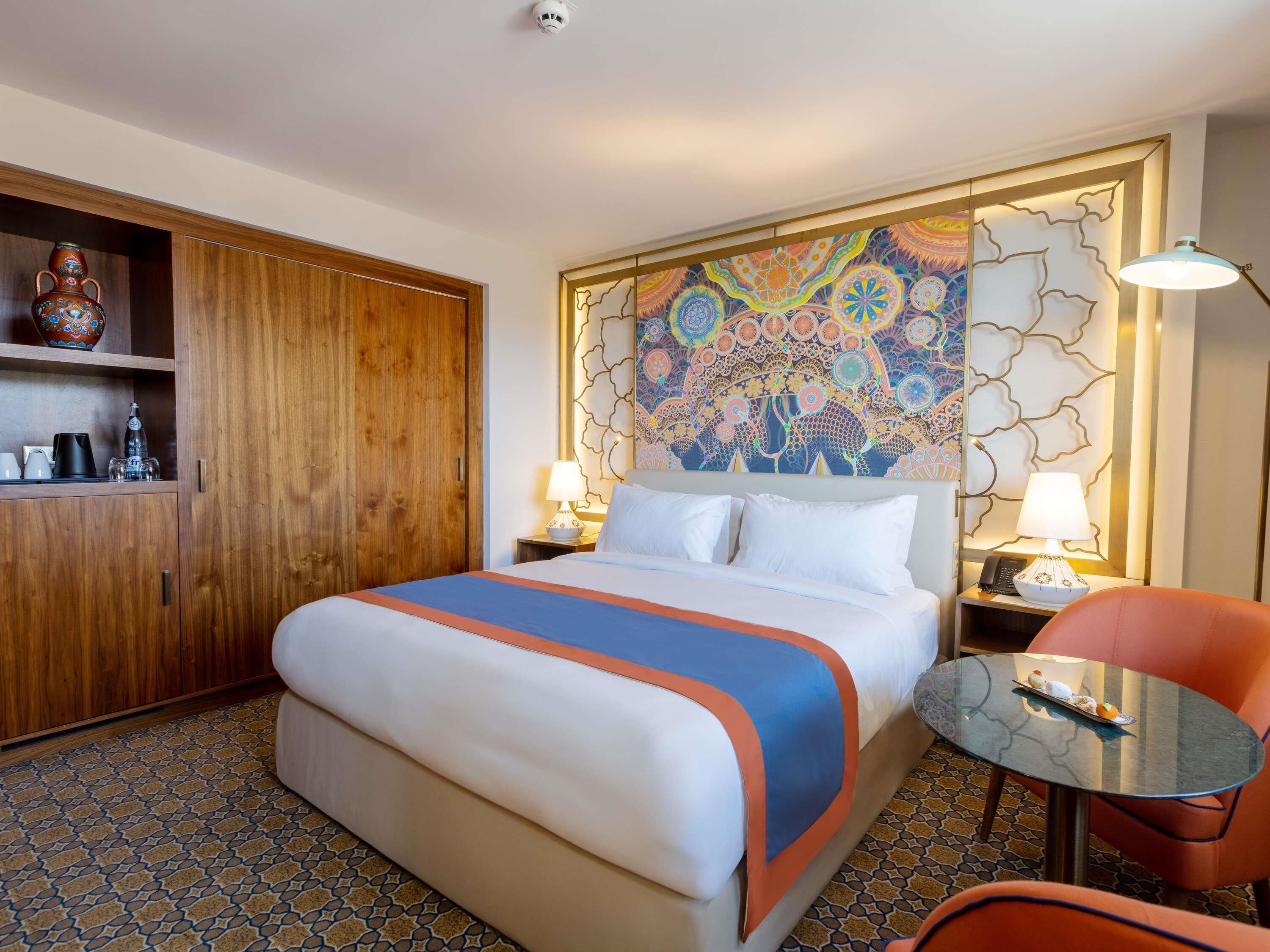 Una habitación de hotel con una cama, un armario de madera, un muro decorativo, una pequeña mesa con una silla y una mesita de noche con una lámpara.