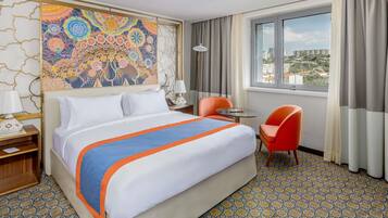 Habitación de hotel con una cama grande, un escritorio con silla, una ventana con vistas a la ciudad y un cuadro abstracto colorido en la pared.