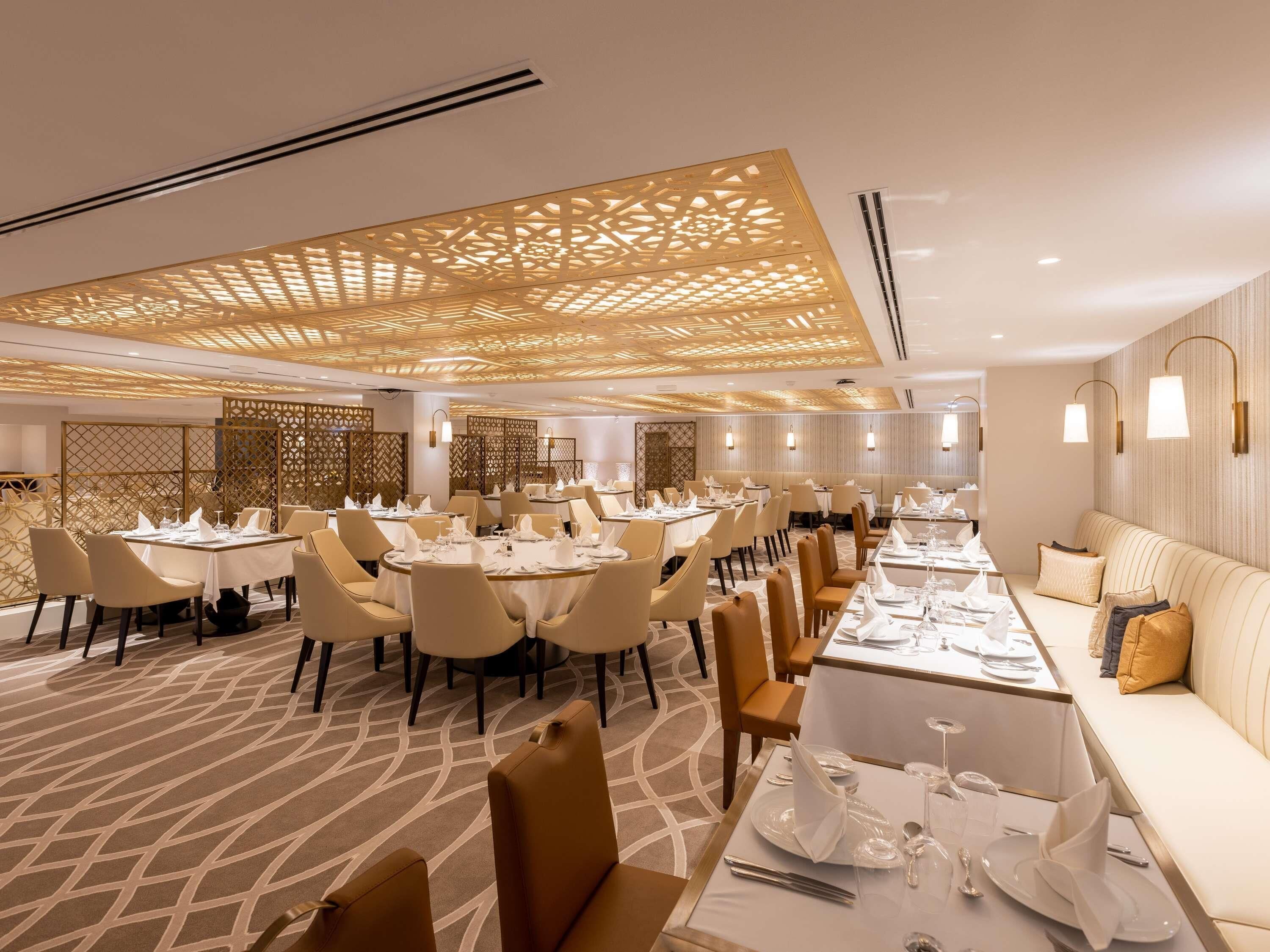Un área de comedor moderna con mesas y sillas redondas, una barra alargada con sofá y paneles decorativos en el techo.