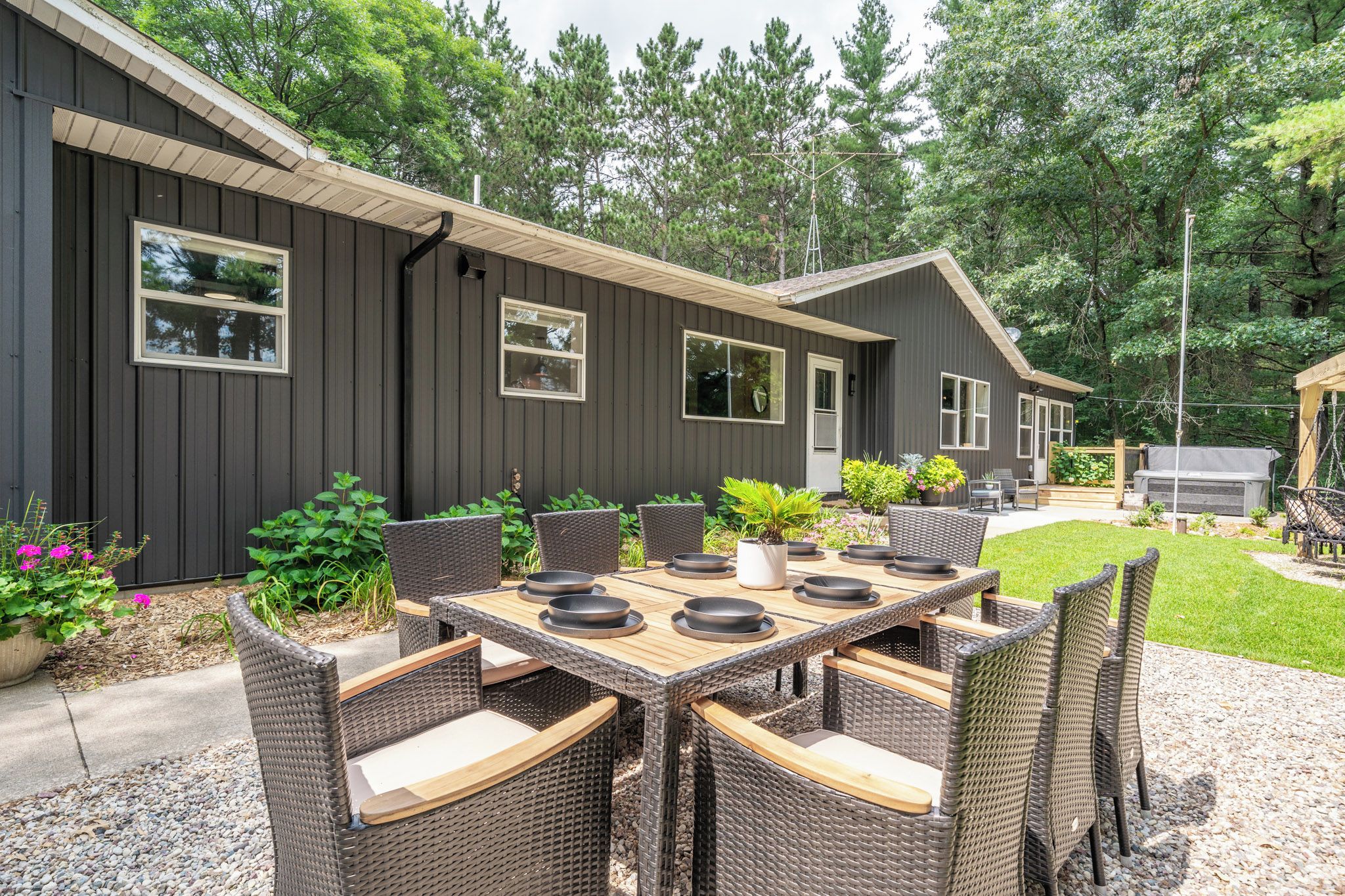 A modern outdoor dining area with a large table and wicker chairs, surrounded by a well-maintained garden and a dark-colored building.