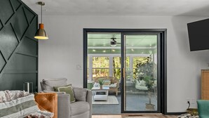 Ein modernes Wohnzimmer mit Sofa, Sessel und Couchtisch. Eine Schiebetür aus Glas führt zu einem Außenbereich mit Stuhl und Tisch.