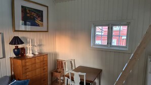 Une pièce comprenant une commode en bois, une table, une chaise, une lampe, un tableau encadré accroché au mur et une fenêtre donnant sur des bâtiments rouges.