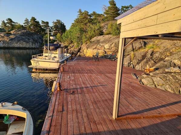 A wooden dock with chairs and a boat docked at the edge of a calm lake, surrounded by trees and rocky terrain.