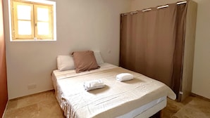A neatly made bed with a white comforter and two pillows, a window with a wooden frame, and a brown curtain.