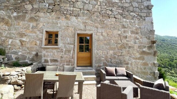 A stone building with a wooden door and two windows, outdoor wicker furniture, and a view of the surrounding landscape.