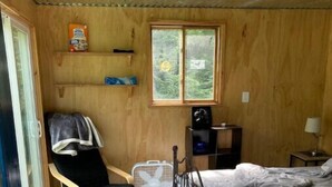 Una stanza compatta con un letto, una sedia, un tavolino, una finestra e uno scaffale con oggetti.