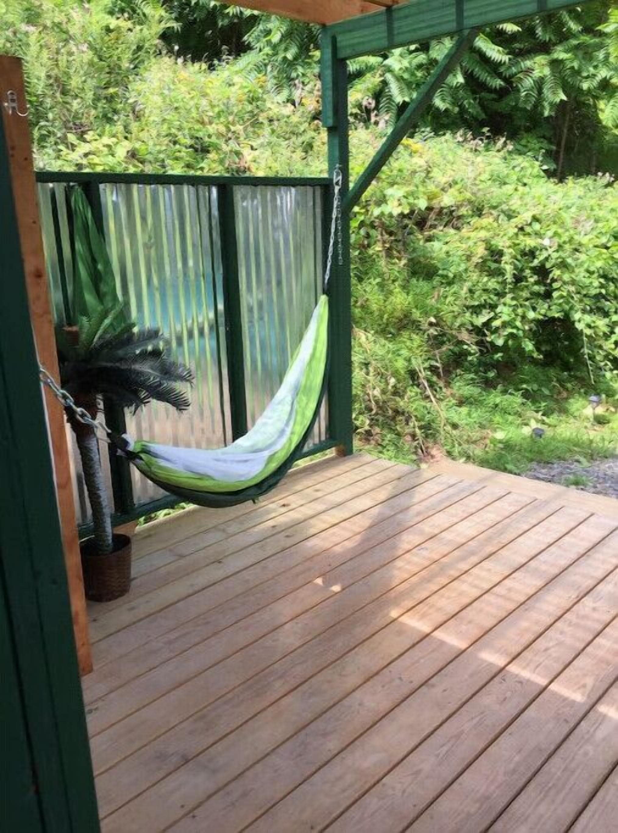 A hammock hanging on a wooden deck with a metal railing and a green structure in the background.