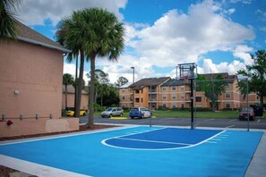 Ein Basketballplatz neben einem Gebäude mit einem Parkplatz und einem Wohnkomplex im Hintergrund.