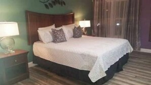A neatly made bed with a white bedspread and multiple pillows, a wooden nightstand with a lamp, and a window with curtains. - Splash of Disney Color & Fun (Kissimmee)