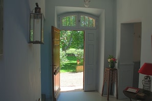 Een hal met een houten deur die uitkomt op een tuin. Aan de rechterkant staat een kleine tafel met een bloemenvaas en een lamp.