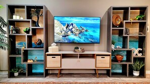 A modern living room with a large flat-screen TV mounted on the wall, wooden shelving units on either side, and a central desk with two drawers.