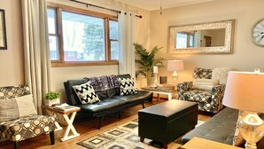 A living room with a sofa, ottoman, and chairs, a coffee table, a lamp, a mirror, and a clock on the wall.