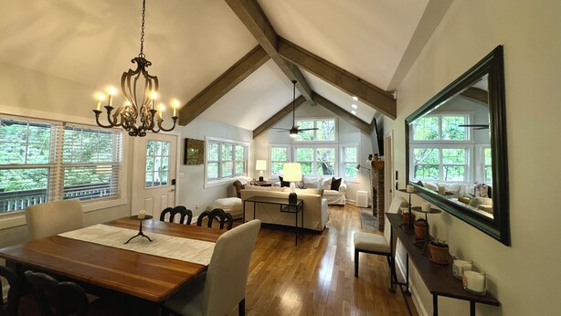 A spacious living area with a wooden dining table, comfortable seating, a chandelier, and large windows offering views of greenery.