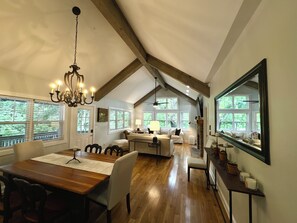 A spacious living area with a wooden dining table, comfortable seating, a chandelier, and large windows offering views of greenery. - Chestnut Cottage (Highlands)