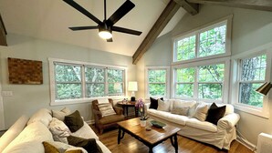 A spacious living room with a wooden floor, two white sofas, a brown armchair, a wooden coffee table, and a ceiling fan. Large windows provide natural light and views of greenery outside.
