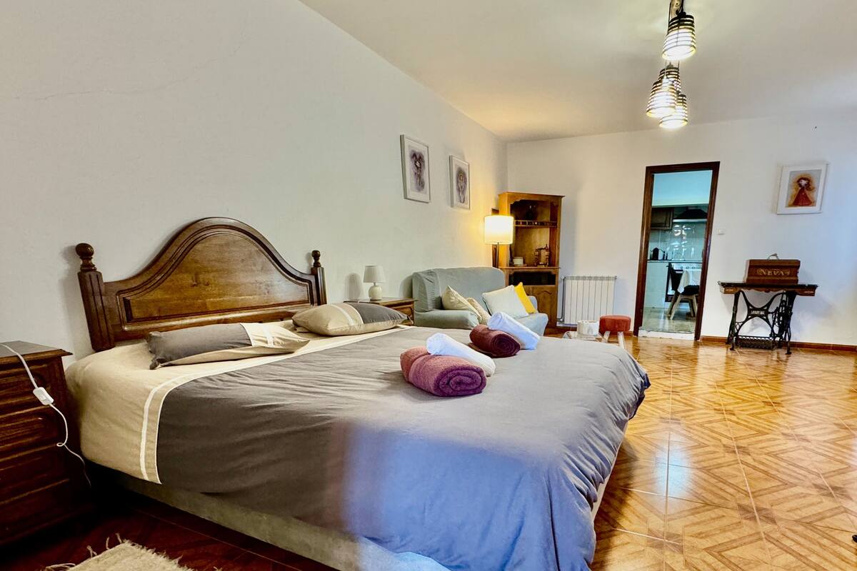 A neatly made bed with a wooden headboard, a bedside table with a lamp, and a chair in a room with a herringbone floor pattern.