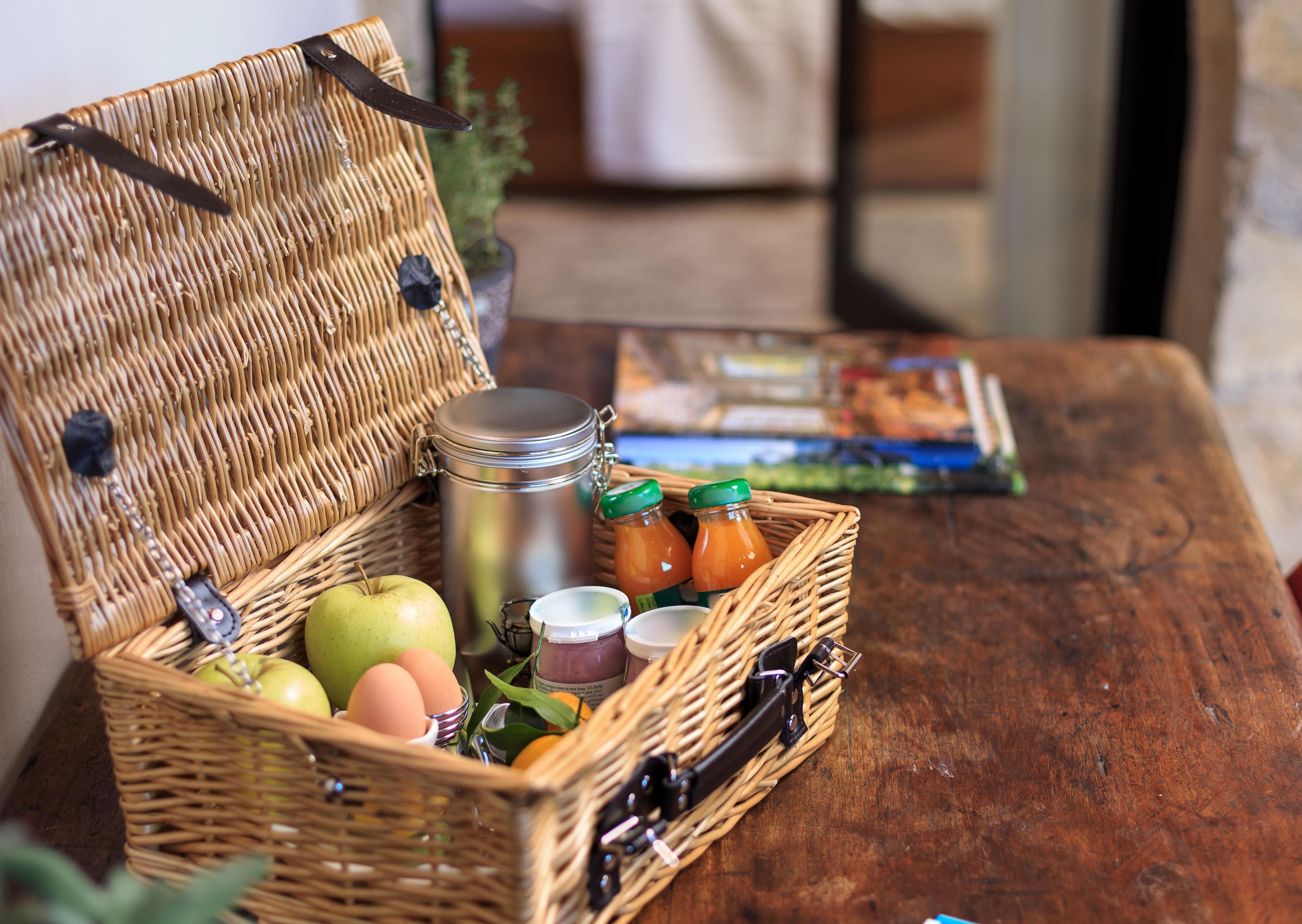A wicker basket with a metal container, jars, an apple, and eggs on a wooden table.