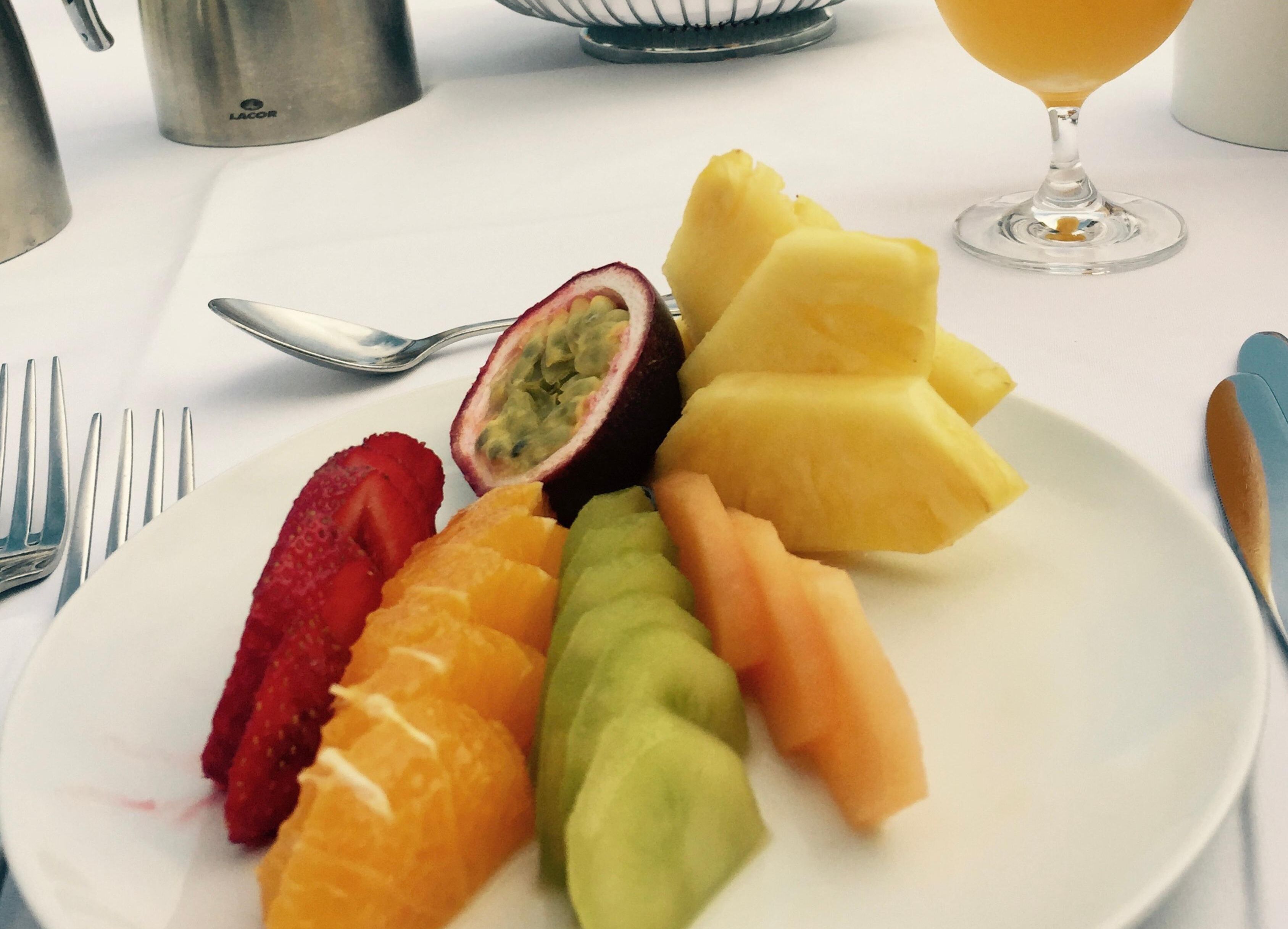 Un plato de frutas cortadas, incluyendo fresas, kiwi y melón, con un vaso de jugo de naranja al fondo.