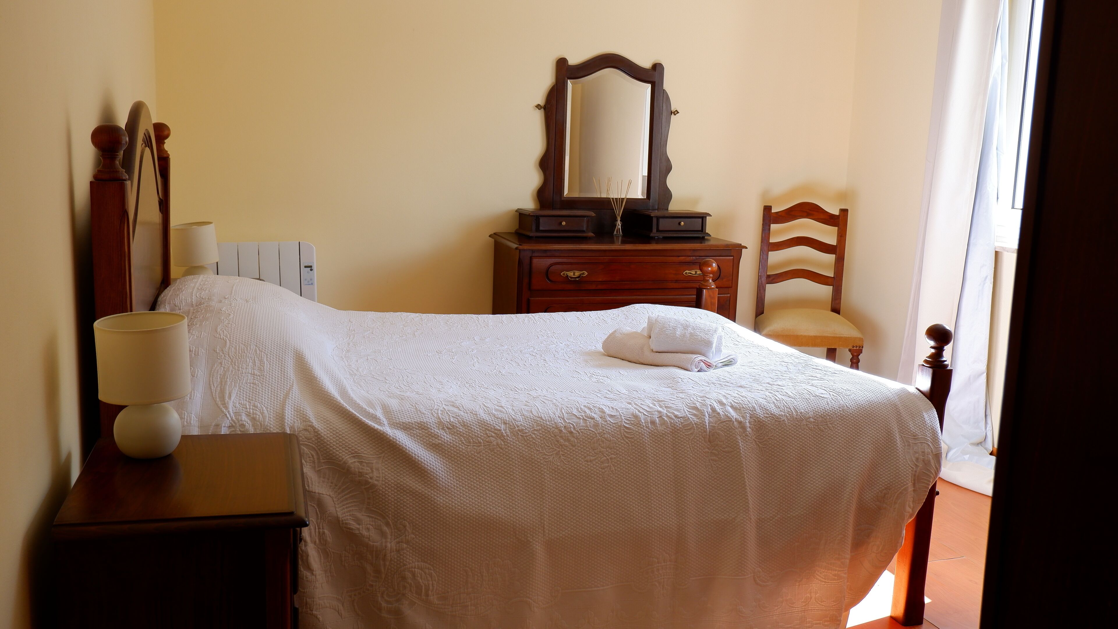 A neatly made bed with a white bedspread, a wooden dresser with a mirror, a chair, and a bedside table with a lamp.