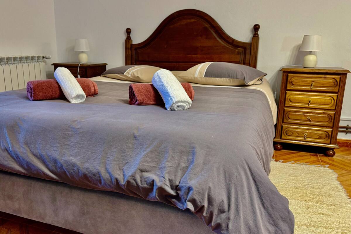 A neatly made bed with a grey bedspread and multiple towels, a wooden headboard, a nightstand with a lamp, and a dresser with drawers.