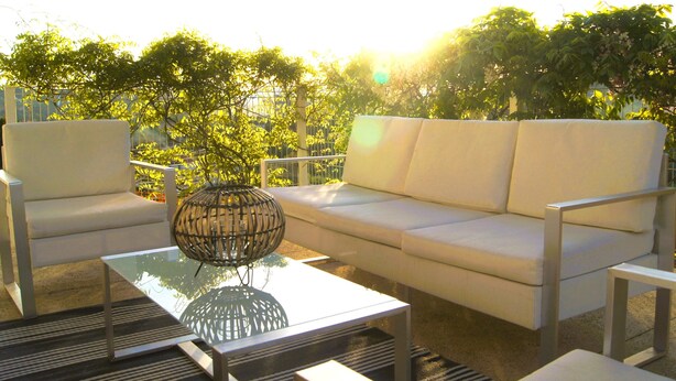 Un área de estar exterior moderna con un sofá blanco, dos sillones blancos y una mesa de centro de vidrio con una lámpara trenzada.