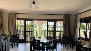 A dining area with a large table and chairs, large windows with metal grilles, and a ceiling fan. - The Fortuna Hotel and Cafe (Kabale)