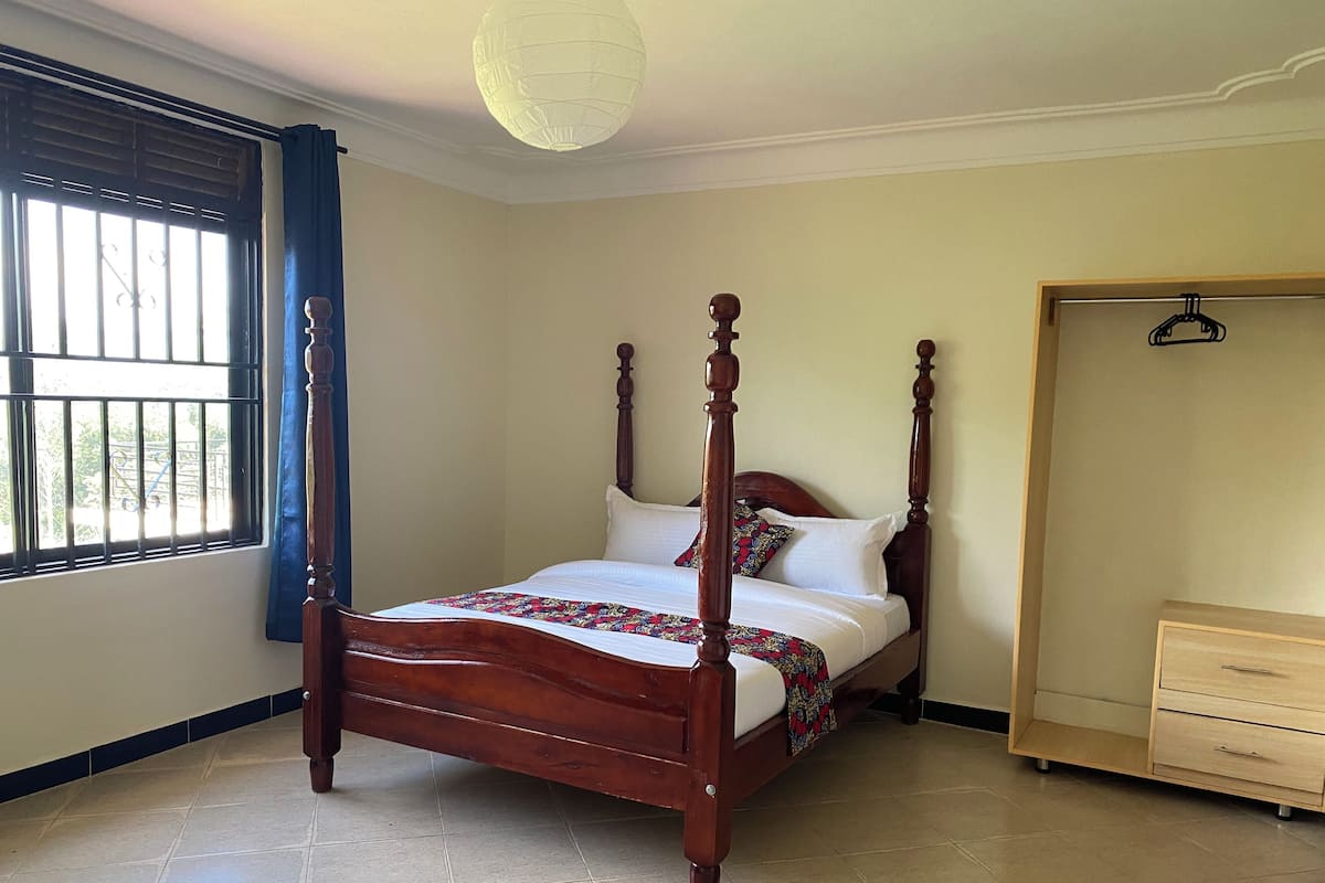 A four-poster bed with a wooden frame, a white bedspread, and a colorful throw blanket. There is a wooden wardrobe with drawers to the right of the bed.