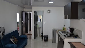 A compact living space with a kitchenette, a blue sofa, a small dining table, and a fan. - Casa Confort (Pereira)