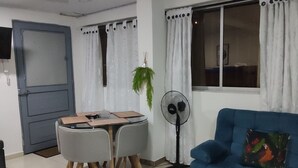 A living room with a blue sofa, a dining table with chairs, a standing fan, and a door. - Casa Confort (Pereira)