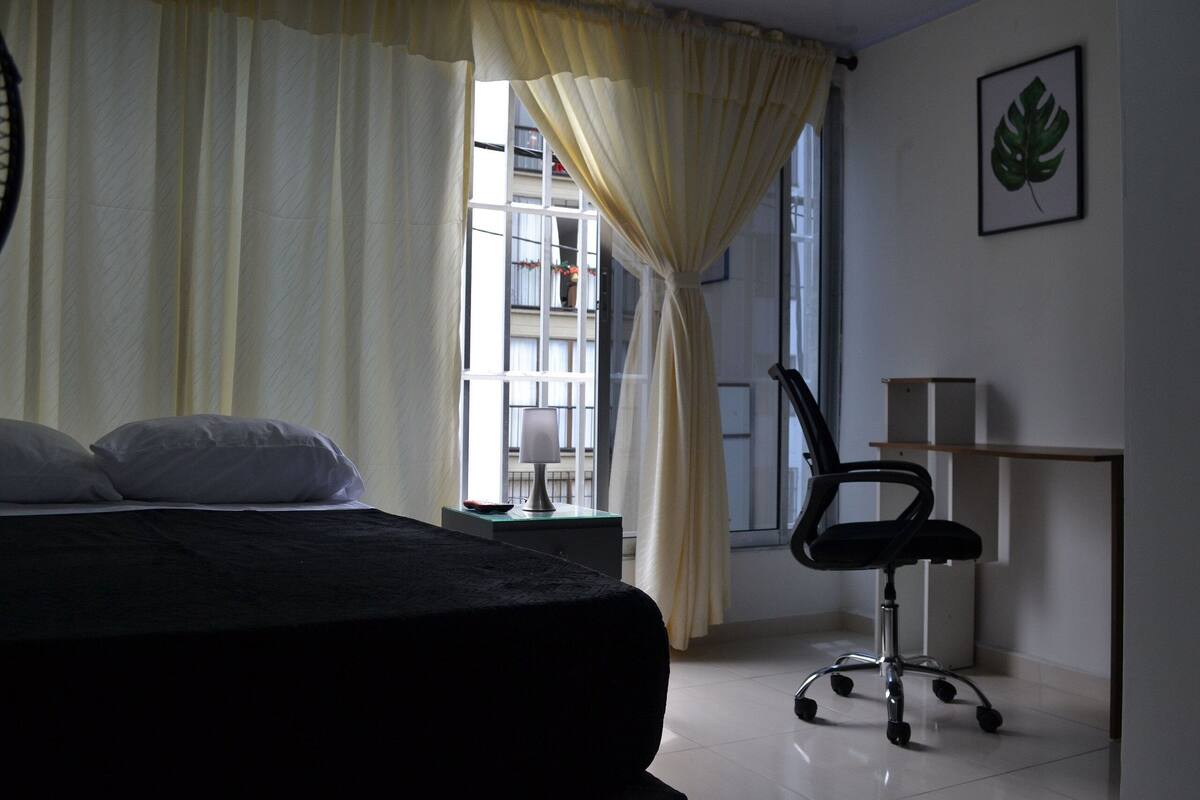 A bedroom with a bed, a desk, and a chair. There is a window with curtains and a plant on the wall.