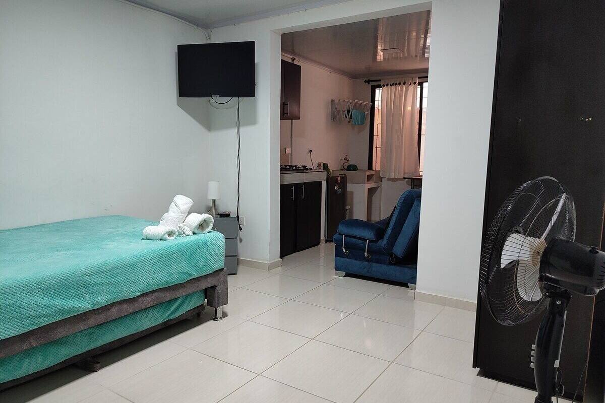 A room with a bed, a TV mounted on the wall, a fan, and a kitchenette area in the background.