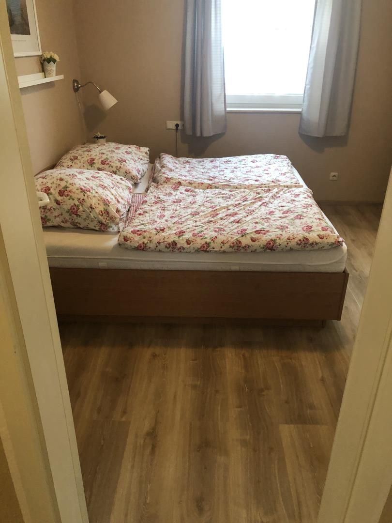 A bedroom with a double bed, a bedside lamp, a window with curtains, and a wooden floor.