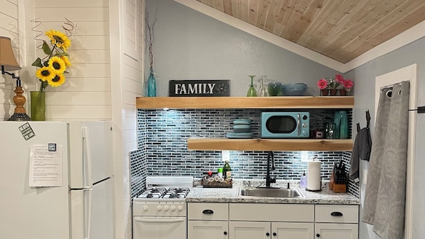 Une cuisine de taille réduite avec des meubles blancs, un four à micro-ondes, une cuisinière et un évier. Un panneau indiquant « FAMILLE » est accroché au mur, un vase avec des tournesols est posé sur une étagère, et un tapis bleu recouvre le sol.