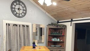 Un coin repas avec une table en bois et quatre chaises, un ventilateur de plafond, une horloge, un miroir et une étagère avec des bouteilles et de la vaisselle.