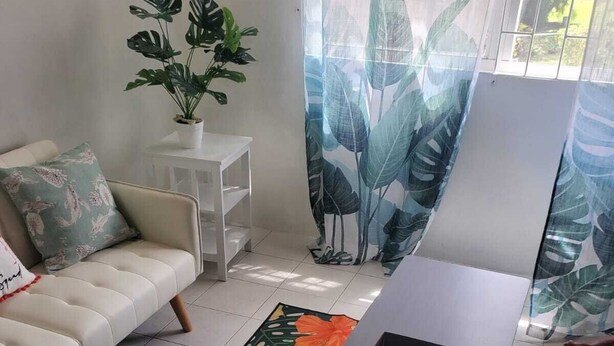 A living room with a sofa, a small white table, a dark wooden coffee table with decorative items, and a large window with curtains. - Tropical Paradise (Bridgetown)